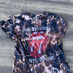 May include: A black and brown tie-dye t-shirt featuring the Rolling Stones' iconic tongue and lips logo in red and blue. The text "THE ROLLING STONES" and "1975 - US TOUR" are printed on the shirt.