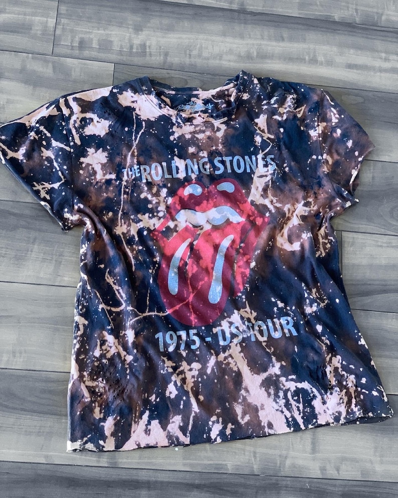 May include: A black and brown tie-dye t-shirt featuring the Rolling Stones' iconic tongue and lips logo in red and blue. The text "THE ROLLING STONES" and "1975 - US TOUR" are printed on the shirt.