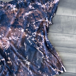 May include: A short-sleeved t-shirt with a unique, abstract design. The shirt features a dark navy blue base color with a bleached, splattered pattern in shades of brown and white, creating a marbled effect. The fabric appears soft and casual.