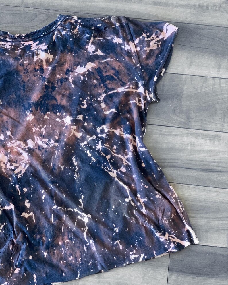 May include: A short-sleeved t-shirt with a unique, abstract design. The shirt features a dark navy blue base color with a bleached, splattered pattern in shades of brown and white, creating a marbled effect. The fabric appears soft and casual.