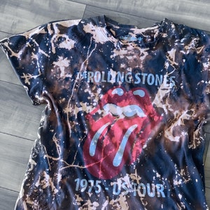 May include: Black t-shirt with a bleach-splattered design. The shirt features the Rolling Stones' iconic red lips logo and the text "THE ROLLING STONES 1975 - US TOUR" in a light blue font. The shirt has a crew neck and short sleeves.