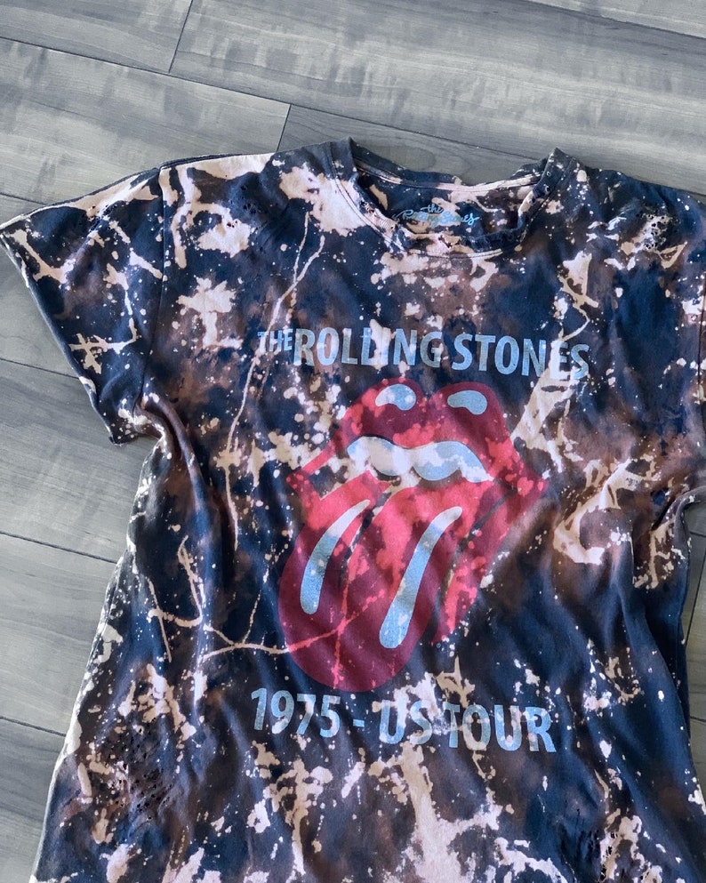 May include: Black t-shirt with a bleach-splattered design. The shirt features the Rolling Stones' iconic red lips logo and the text "THE ROLLING STONES 1975 - US TOUR" in a light blue font. The shirt has a crew neck and short sleeves.