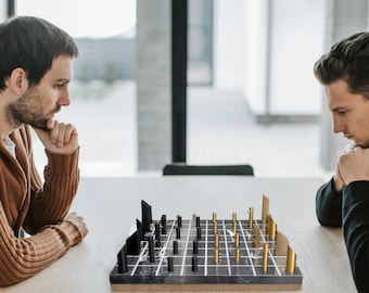 May include: A modern chess set with black and gold pieces on a marble-patterned board. The pieces include stylized towers and cylindrical pieces. Two people are seated at a table, looking at the chess board.