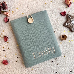 Könnte beinhalten: Hellblau-grüne, gesteppte Buchhülle mit dem Namen "Emily" in passendem Faden gestickt. Sie wird mit einem Holzknopf und einer Schlaufe verschlossen. Der Hintergrund zeigt eine sternförmige Dekoration und florale Akzente.