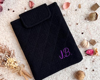 May include: Black quilted tablet case with a flap closure. The case has a diamond pattern and embroidered purple initials "J.B." on the front. The case is on a white, textured surface with floral accents.