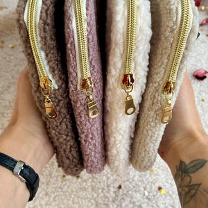 Può includere: Una collezione di quattro pochette soffici con cerniera, nei toni del marrone, malva, bianco e beige. Ogni pochette presenta una cerniera color oro. Le pochette sono realizzate in un materiale morbido e strutturato.