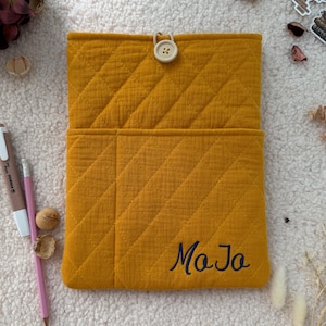 May include: A mustard yellow quilted fabric pouch featuring a diagonal stitched pattern, a single light-coloured button closure at the top, and the name "MoJo" embroidered in dark blue thread on the lower right corner of the front pocket.