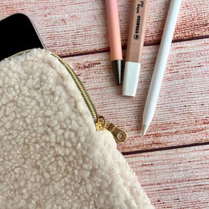 May include: A cream-colored, fluffy pouch with a gold zipper, next to a black tablet, a pink pen, a brown highlighter, and a white stylus. The items are arranged on a wooden surface.