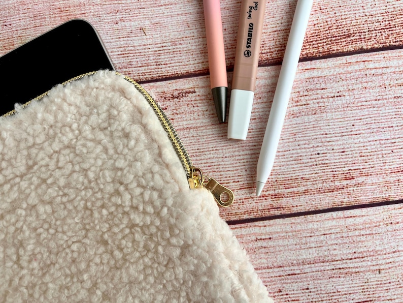 May include: A cream-colored, fluffy pouch with a gold zipper, next to a black tablet, a pink pen, a brown highlighter, and a white stylus. The items are arranged on a wooden surface.