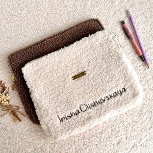 May include: Two fluffy, rectangular pouches, one brown and one cream-colored, are stacked on a textured surface. The cream pouch has the name "Irisha Olishevskaya" embroidered on it, along with a small gold-colored metal tag. A pen and pencil are next to the pouches.
