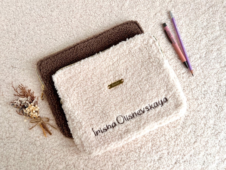 May include: Two fluffy, rectangular pouches, one brown and one cream-colored, are stacked on a textured surface. The cream pouch has the name "Irisha Olishevskaya" embroidered on it, along with a small gold-colored metal tag. A pen and pencil are next to the pouches.