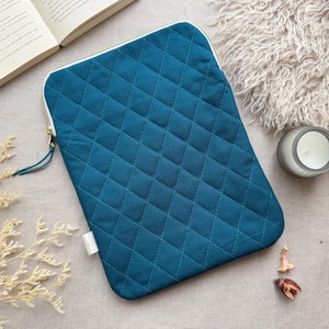 May include: A rectangular teal laptop sleeve featuring a quilted diamond pattern across the front surface. The sleeve has a white zipper closure along the top and side, accented with a matching teal fabric pull tab and a small white side tag.