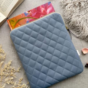 May include: A slate blue quilted laptop sleeve featuring a diamond-stitched pattern and a gold-toned zipper closure. The rectangular protective case has rounded corners and a smooth fabric texture, designed to hold a portable electronic device securely.