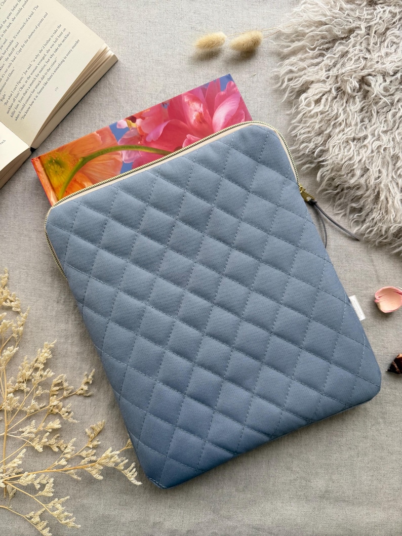 May include: A slate blue quilted laptop sleeve featuring a diamond-stitched pattern and a gold-toned zipper closure. The rectangular protective case has rounded corners and a smooth fabric texture, designed to hold a portable electronic device securely.
