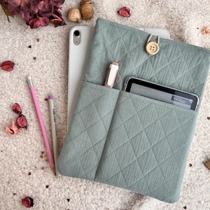 Puede incluir: Una funda acolchada para tableta de color verde salvia con cierre de botón. La funda tiene bolsillos que contienen un lápiz óptico y una tableta pequeña. Una tableta blanca y dos lápices están junto a la funda. El fondo es una superficie suave y blanca con elementos decorativos.
