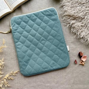 May include: A rectangular, teal-colored laptop sleeve featuring a quilted diamond pattern across the front surface. The sleeve includes a gold-toned metal zipper closure with a matching teal fabric pull tab and a small white brand tag on the side.
