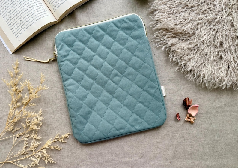 May include: A rectangular, teal-colored laptop sleeve featuring a quilted diamond pattern across the front surface. The sleeve includes a gold-toned metal zipper closure with a matching teal fabric pull tab and a small white brand tag on the side.