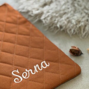 May include: A rectangular, quilted fabric mat in a solid burnt orange color. The surface features a diamond-shaped stitched pattern and the name Serna embroidered in white cursive script across the lower portion of the fabric.