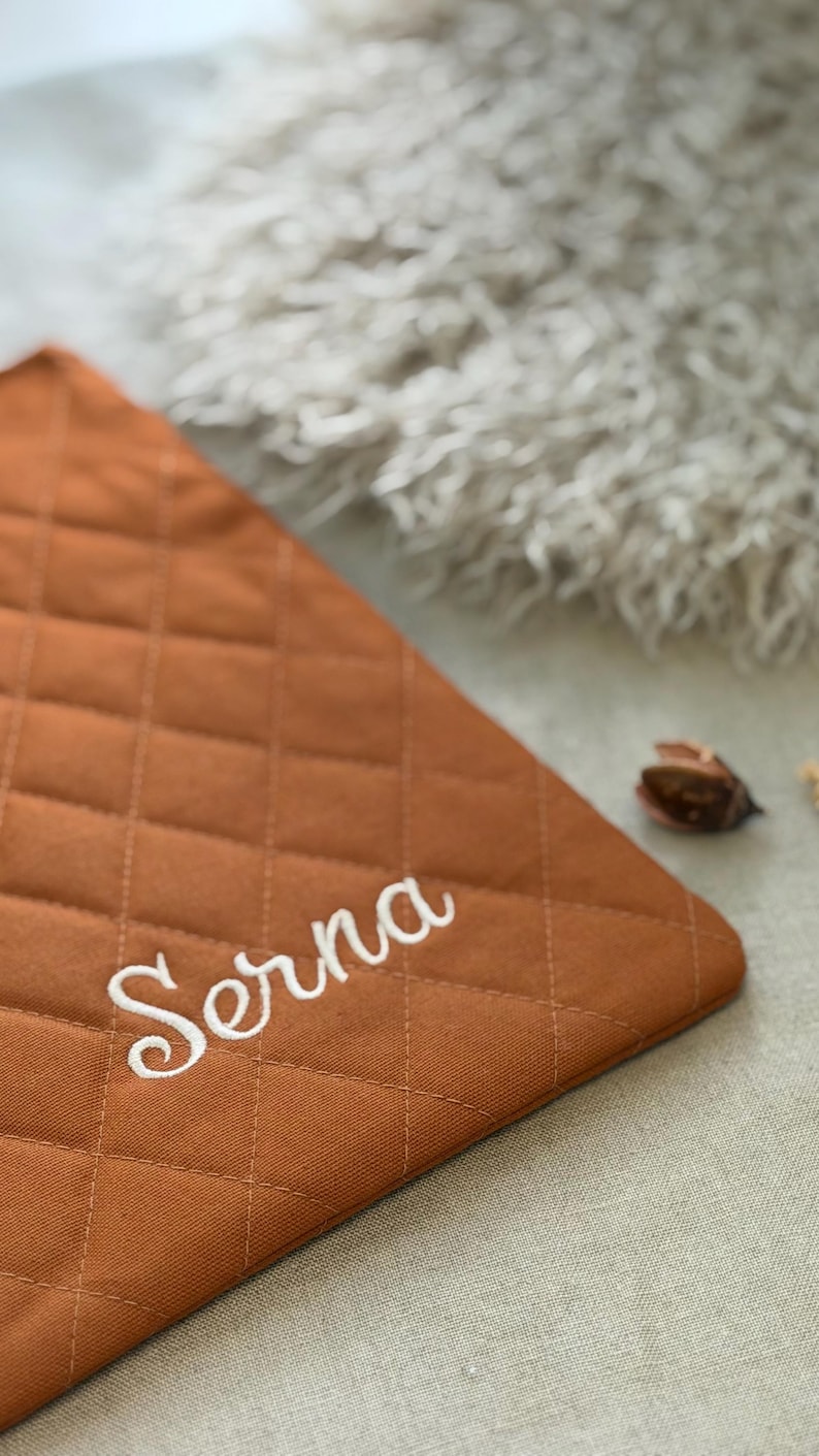 May include: A rectangular, quilted fabric mat in a solid burnt orange color. The surface features a diamond-shaped stitched pattern and the name Serna embroidered in white cursive script across the lower portion of the fabric.