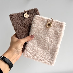 Puede incluir: Dos bolsas rectangulares y esponjosas en colores marrón y crema. Cada bolsa tiene un cierre de botón de madera. Las bolsas están hechas de un material suave y texturizado, y se sostienen en una mano.