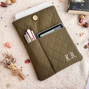 Puede incluir: Funda acolchada para tableta de color verde oliva con bolsillos para bolígrafos y un teléfono. La funda tiene un cierre de botón de madera y las iniciales "K.R." bordadas en la parte delantera. La funda está abierta, revelando una tableta y bolígrafos.
