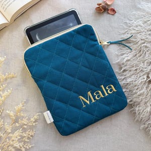 May include: A teal quilted tablet sleeve featuring a diamond-stitched pattern and a gold zipper closure. The name Mala is embroidered in gold thread on the front. A small white fabric tag is attached to the side of the rectangular protective case.