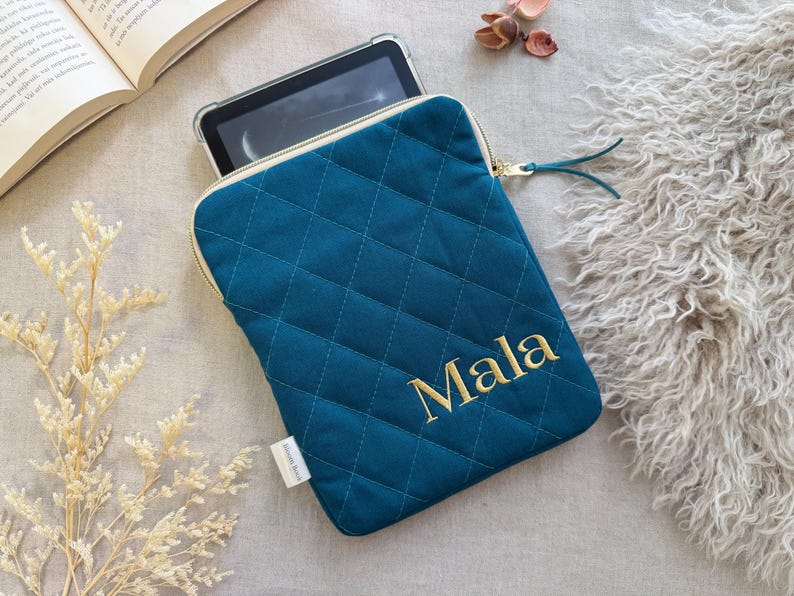 May include: A teal quilted tablet sleeve featuring a diamond-stitched pattern and a gold zipper closure. The name Mala is embroidered in gold thread on the front. A small white fabric tag is attached to the side of the rectangular protective case.