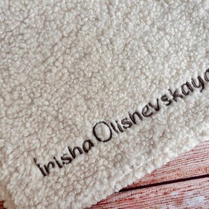 May include: Cream-colored, plush blanket with the embroidered name "Irisha Olishevskaya" in dark brown thread. The blanket has a textured, fuzzy surface and is likely made of a soft material. The background is a wooden surface.