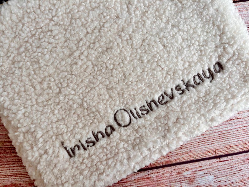 May include: Cream-colored, plush blanket with the embroidered name "Irisha Olishevskaya" in dark brown thread. The blanket has a textured, fuzzy surface and is likely made of a soft material. The background is a wooden surface.
