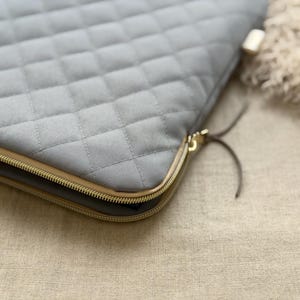May include: A light gray quilted laptop sleeve featuring a diamond-stitched pattern, a gold-toned metal zipper closure, and thin dark gray leather-like zipper pulls. The sleeve has a structured rectangular shape with rounded corners.
