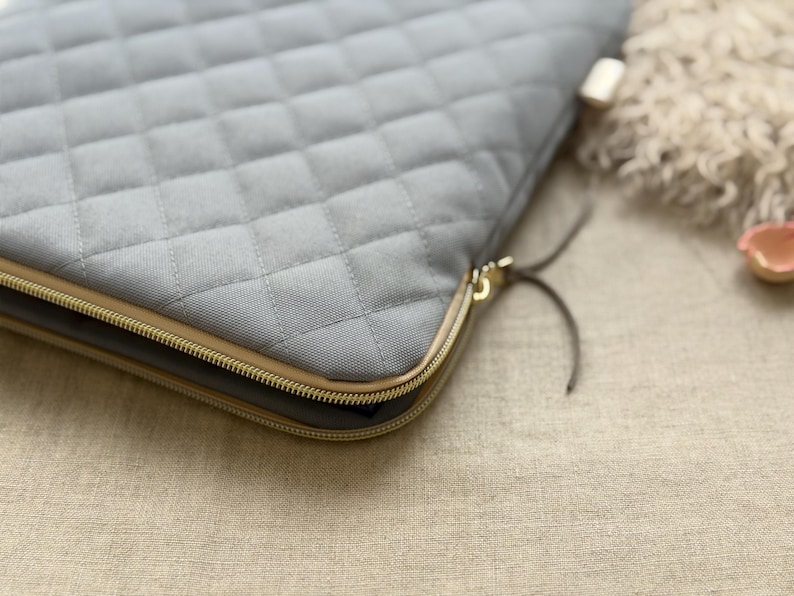 May include: A light gray quilted laptop sleeve featuring a diamond-stitched pattern, a gold-toned metal zipper closure, and thin dark gray leather-like zipper pulls. The sleeve has a structured rectangular shape with rounded corners.