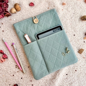 May include: A quilted, seafoam green tablet sleeve with a wooden button closure. The sleeve has two pockets, one holding a pen and the other a tablet. A pink pencil, a gray pencil, and gold paperclips are also visible.