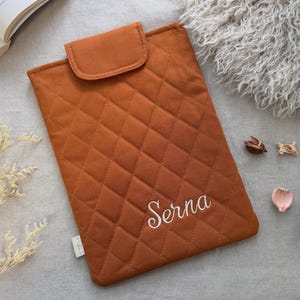 May include: A rectangular, quilted, burnt orange laptop sleeve featuring a flap closure and the name Serna embroidered in white script on the front. The sleeve has a diamond-stitched pattern and a small white fabric tag on the side edge.