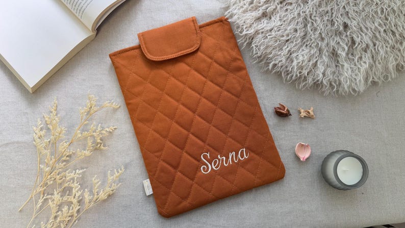 May include: A rectangular, quilted, burnt orange laptop sleeve featuring a flap closure and the name Serna embroidered in white script on the front. The sleeve has a diamond-stitched pattern and a small white fabric tag on the side edge.