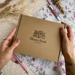 May include: A brown cardboard box with the text "Bloom Book Covers" and a floral illustration is held by a person. The box is on a floral patterned paper with pencils and dried flowers.