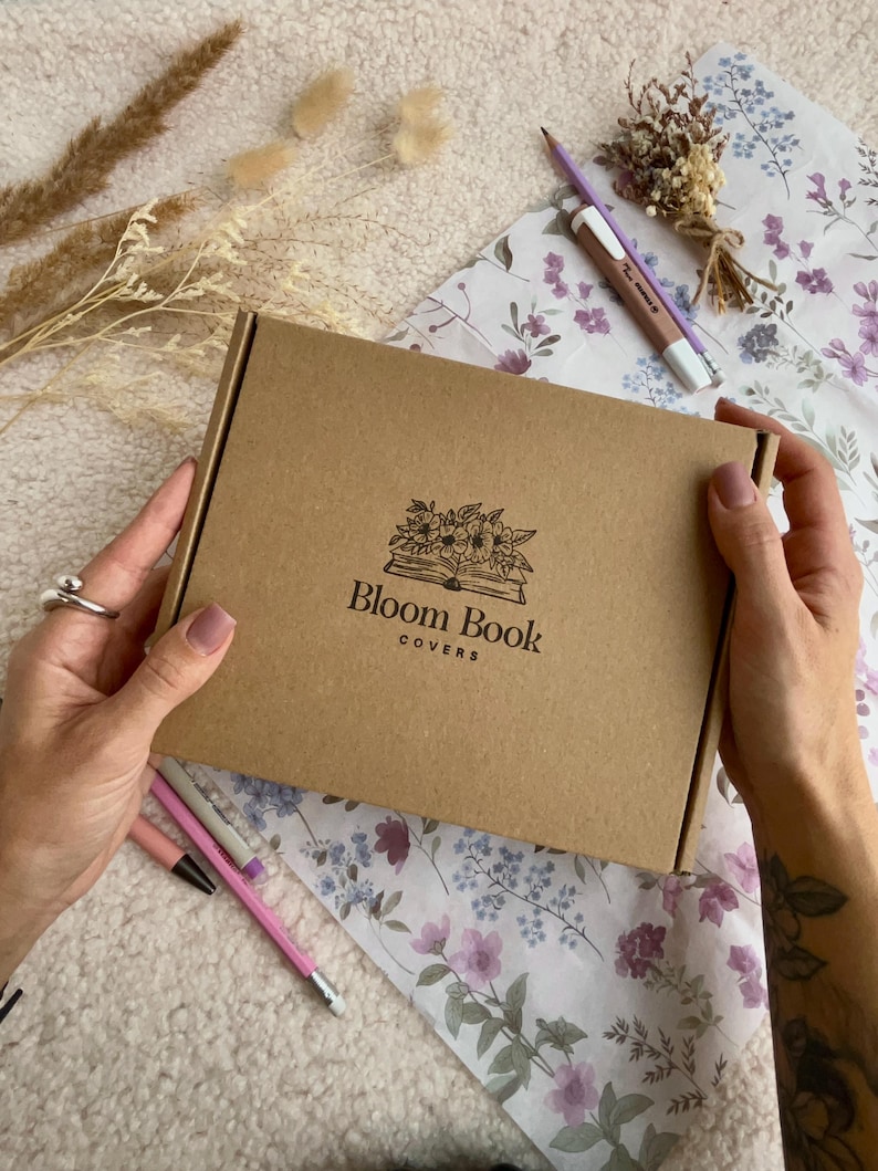 May include: A brown cardboard box with the text "Bloom Book Covers" and a floral illustration is held by a person. The box is on a floral patterned paper with pencils and dried flowers.