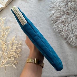 May include: A teal quilted fabric sleeve with a diamond-stitched pattern and a light-colored zipper closure. The rectangular case features a slim profile and a textured exterior, designed to hold flat items securely within its zippered compartment.