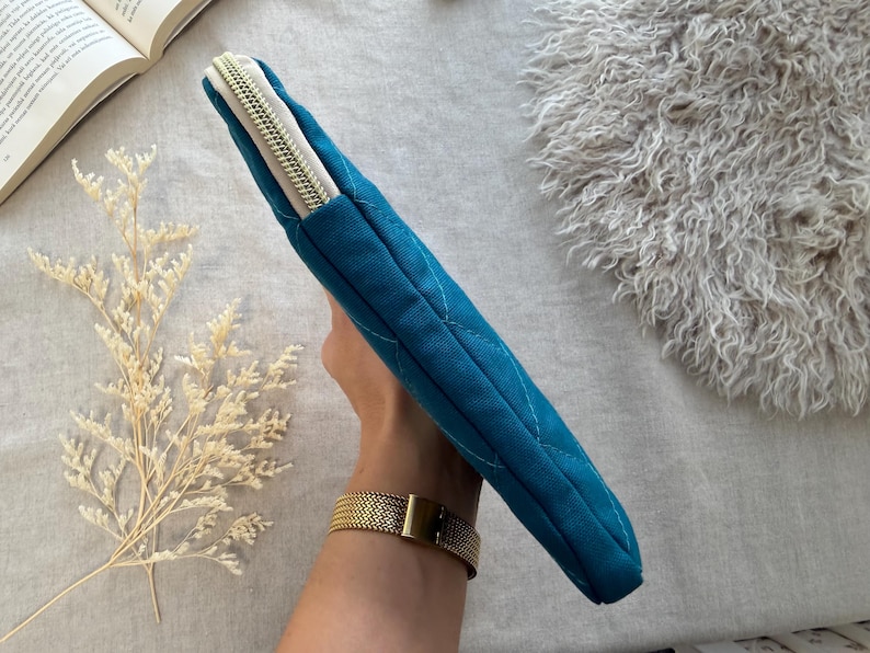 May include: A teal quilted fabric sleeve with a diamond-stitched pattern and a light-colored zipper closure. The rectangular case features a slim profile and a textured exterior, designed to hold flat items securely within its zippered compartment.