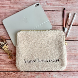 May include: Cream-colored, fluffy tablet sleeve with the embroidered name "Irisha Olishevskaya". A white tablet, pens, and a small bouquet of dried flowers are arranged on a wooden surface.
