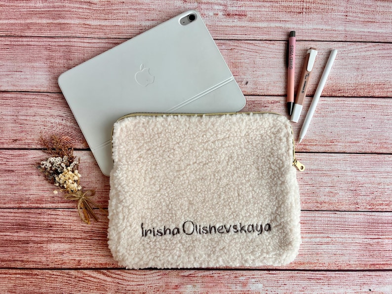May include: Cream-colored, fluffy tablet sleeve with the embroidered name "Irisha Olishevskaya". A white tablet, pens, and a small bouquet of dried flowers are arranged on a wooden surface.