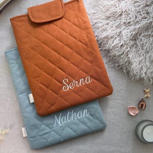 May include: Two rectangular quilted laptop sleeves with flap closures. The top sleeve is burnt orange with the name Serna embroidered in white. The bottom sleeve is dusty blue with the name Nathan embroidered in white. Both feature a diamond-stitched pattern.