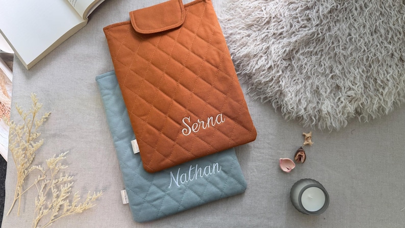 May include: Two rectangular quilted laptop sleeves with flap closures. The top sleeve is burnt orange with the name Serna embroidered in white. The bottom sleeve is dusty blue with the name Nathan embroidered in white. Both feature a diamond-stitched pattern.