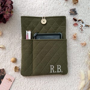 May include: An olive green quilted fabric sleeve featuring a button closure, a front pocket containing a tablet and two pens, and the embroidered initials R.B. in white thread on the bottom right corner of the rectangular protective case.
