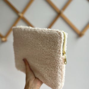 May include: A cream-colored, rectangular clutch made of a soft, textured material. The clutch features a gold-colored zipper closure. The bag is held in a person's hand, showcasing its size and texture.