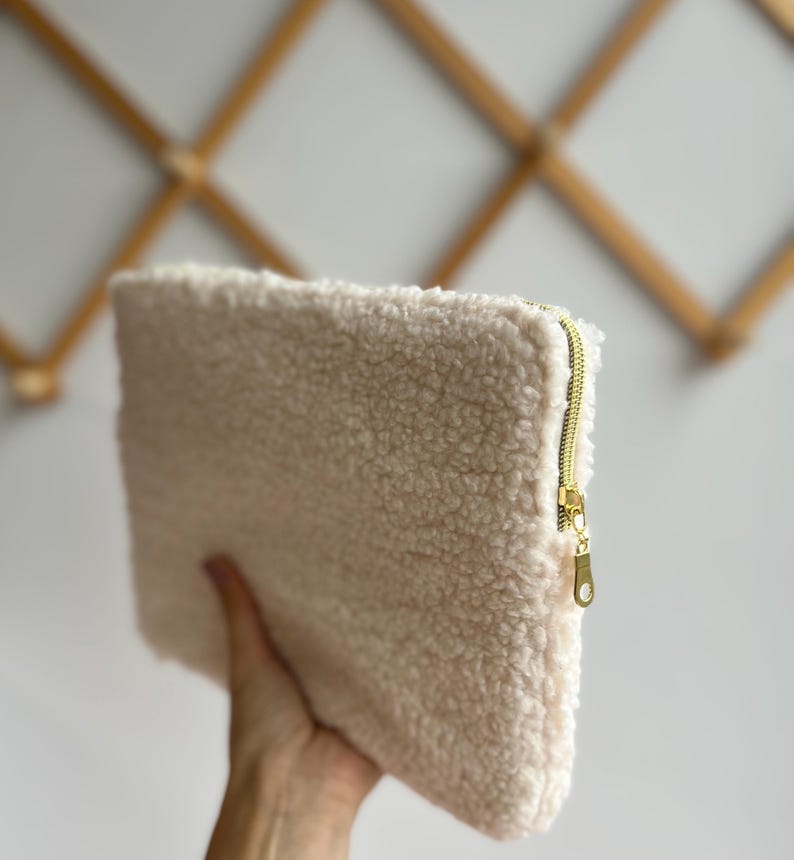 May include: A cream-colored, rectangular clutch made of a soft, textured material. The clutch features a gold-colored zipper closure. The bag is held in a person's hand, showcasing its size and texture.