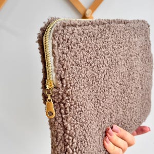 May include: A rectangular, taupe-colored pouch with a textured, fluffy exterior. The pouch features a gold-colored zipper closure. The zipper pull is also gold. The pouch is held by a person.