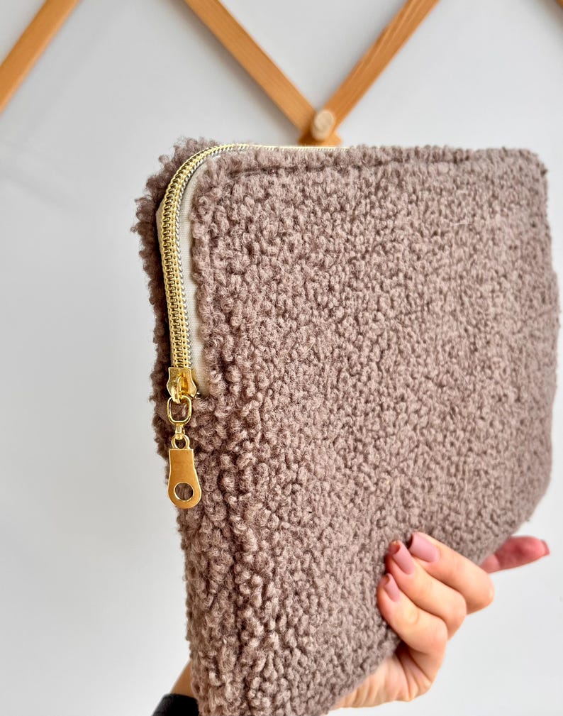 May include: A rectangular, taupe-colored pouch with a textured, fluffy exterior. The pouch features a gold-colored zipper closure. The zipper pull is also gold. The pouch is held by a person.