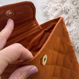 May include: A burnt orange quilted fabric pouch featuring a diamond-stitched pattern and a gold-toned magnetic snap closure. The interior is lined with matching orange fabric, and the exterior flap is secured by a circular metal fastener.