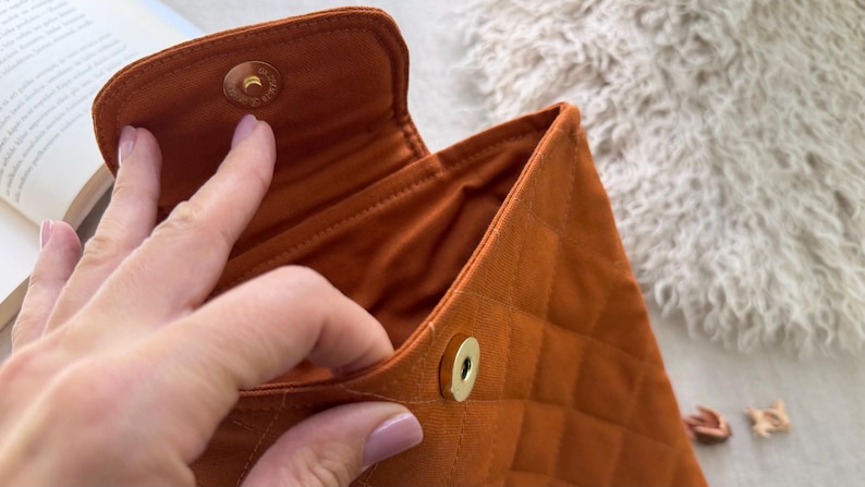 May include: A burnt orange quilted fabric pouch featuring a diamond-stitched pattern and a gold-toned magnetic snap closure. The interior is lined with matching orange fabric, and the exterior flap is secured by a circular metal fastener.