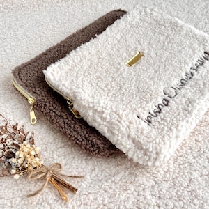 May include: Two fluffy, zippered pouches in brown and white, with gold-toned zippers and a small gold tag. The white pouch has embroidered text. A small bundle of dried flowers rests nearby.
